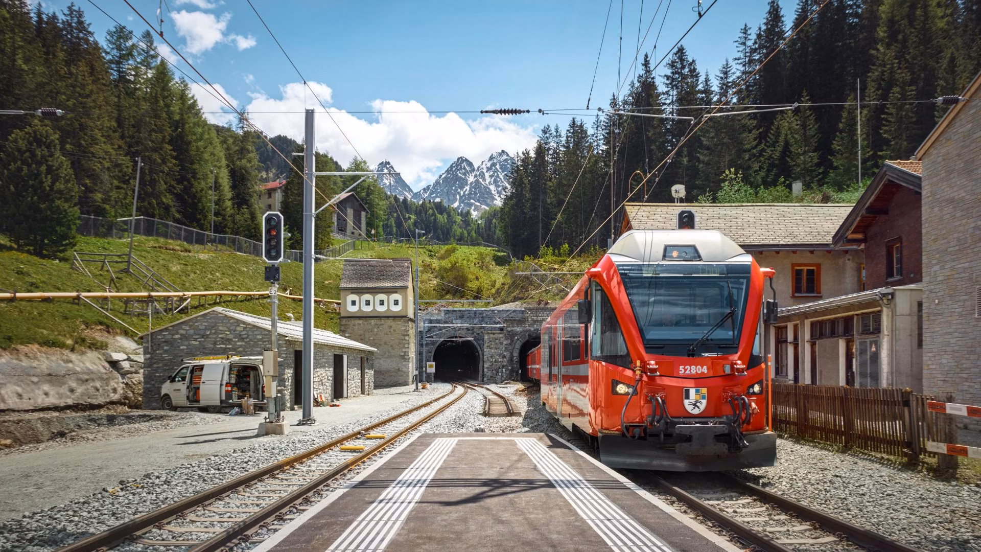 Roter Zug an einem Bergbahnhof mit Gleisen, Tunnel und bewaldeten Alpen im Hintergrund