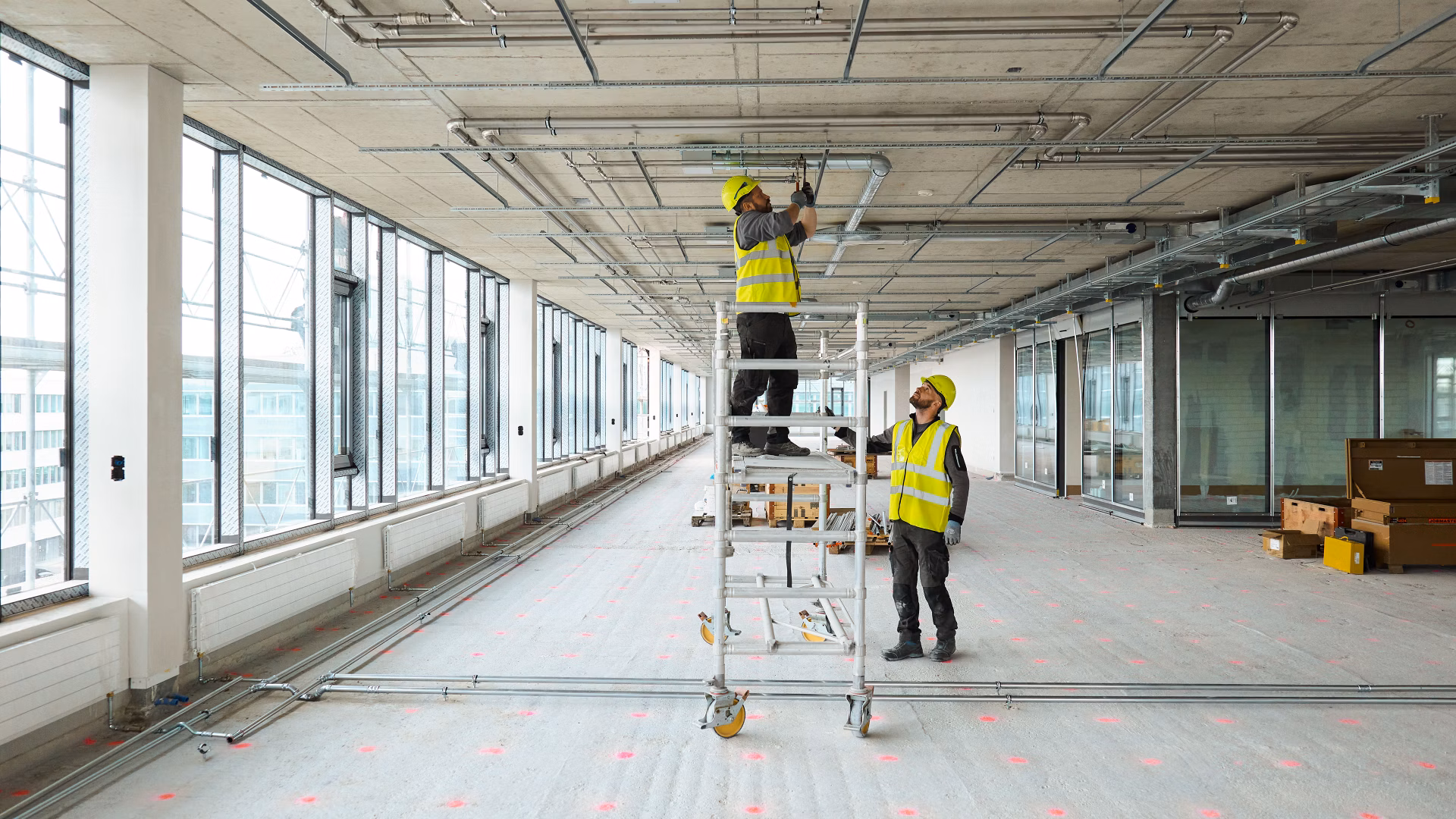 Zwei Installateure arbeiten in einem leeren Innenraum auf einer mobilen Arbeitsplattform an Leitungen an der Decke.