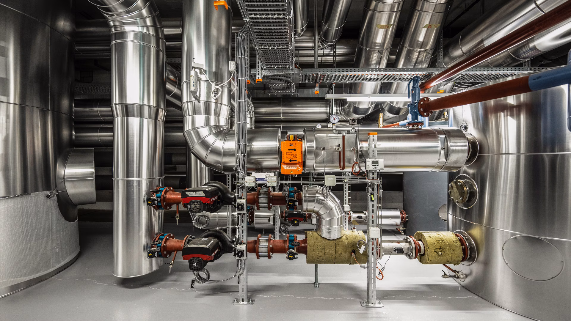 Technikraum mit silbernen Rohrleitungen, Pumpen und grossen Metalltanks eines Wärmeverbunds