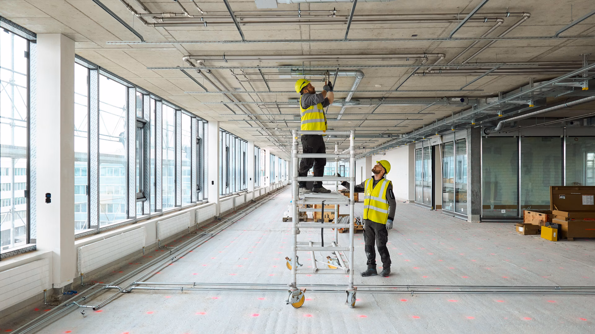 Two workers in high-vis vests install ceiling services in a large unfinished office space.