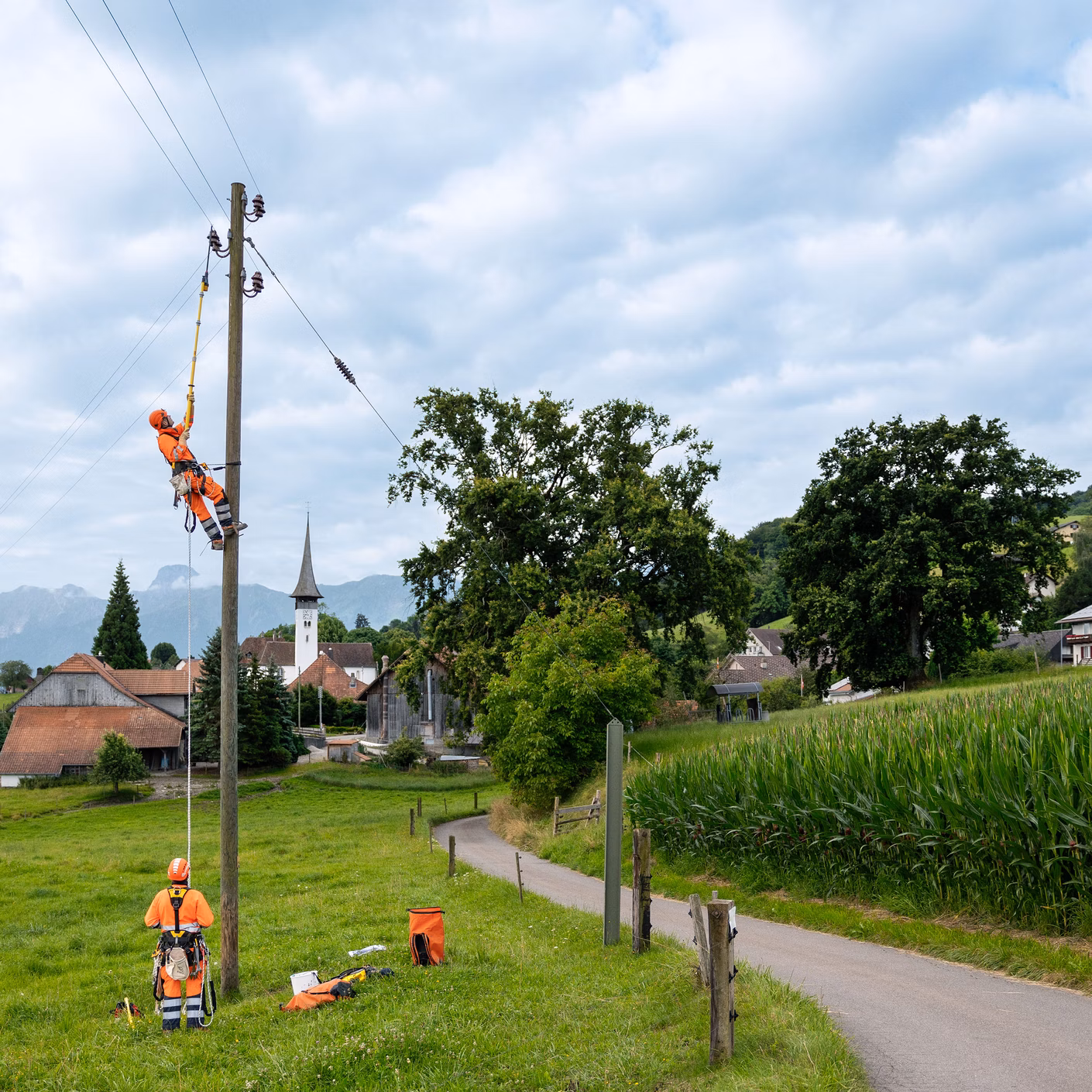 Mitarbeitende arbeiten an einem Strommast neben Feldern und einem Weg in ländlicher Umgebung.