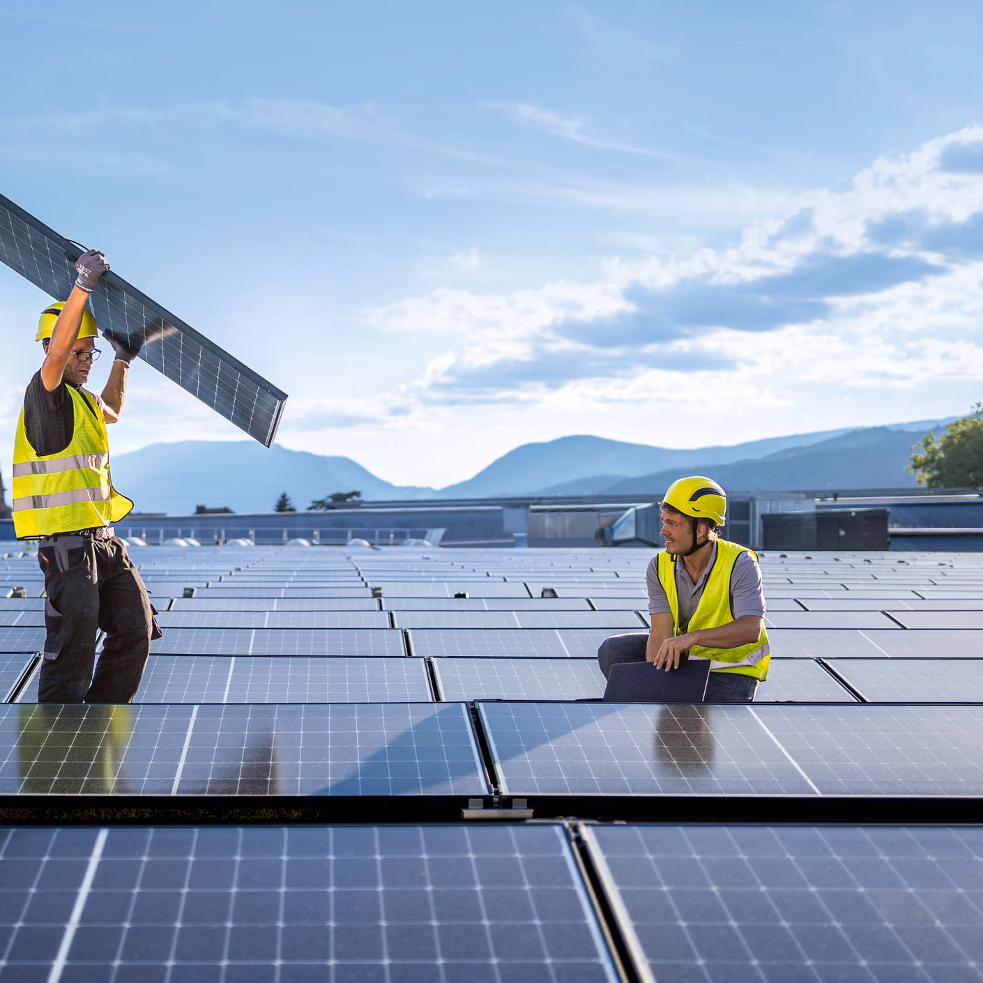 Zwei Arbeiter mit Warnwesten montieren Solarmodule auf einem Dach vor Berglandschaft.