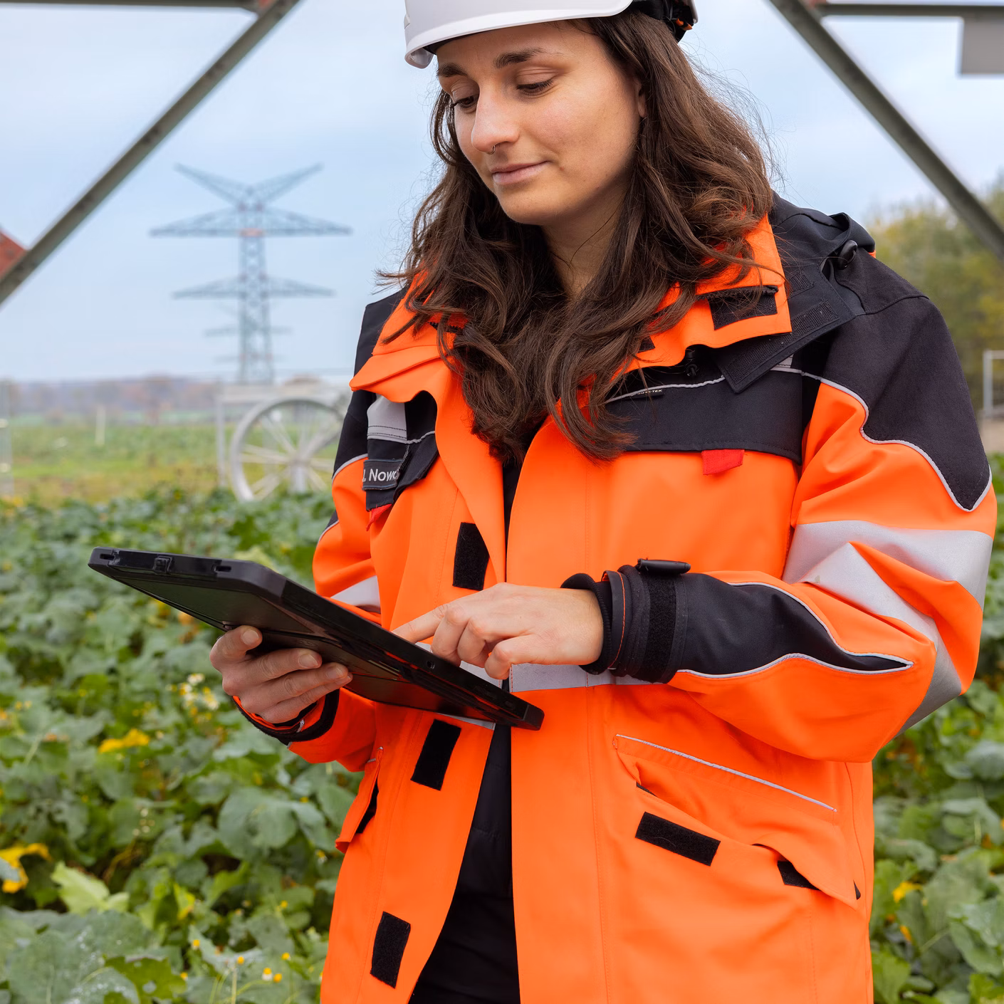 Mitarbeiterin mit Schutzhelm und orangefarbener Arbeitsjacke bedient ein Tablet in einem Gewächshaus.