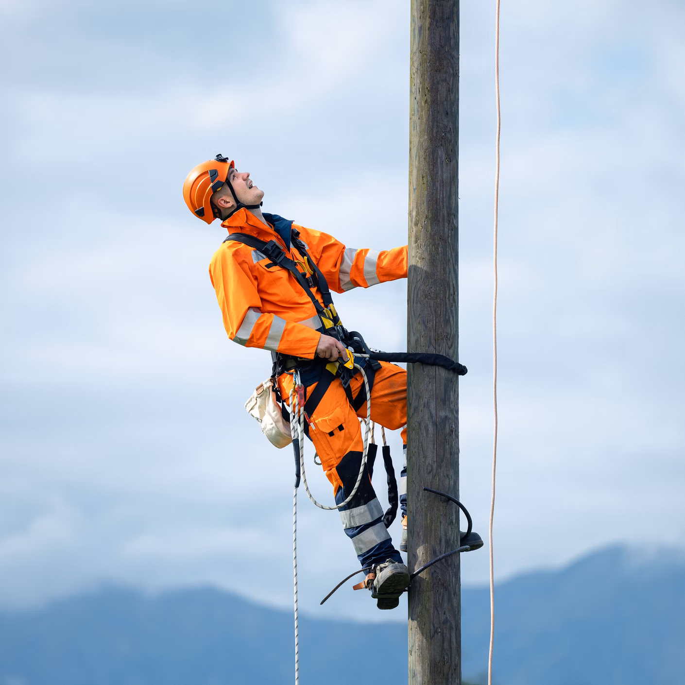 Arbeiter in orangefarbener Schutzkleidung klettert mit Seilsicherung an einem Holzmast.