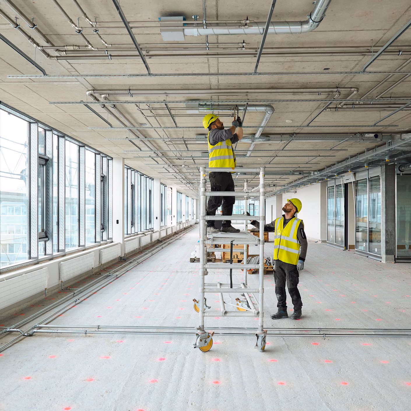 Two workers in high-vis vests install ceiling services in a large unfinished office space.