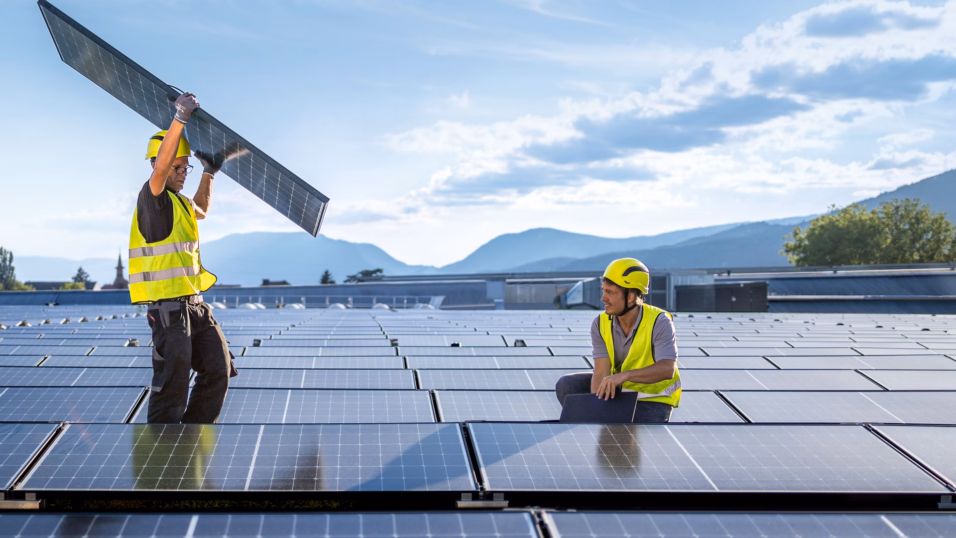 Zwei Arbeiter mit Warnwesten montieren Solarmodule auf einem Dach vor Berglandschaft.