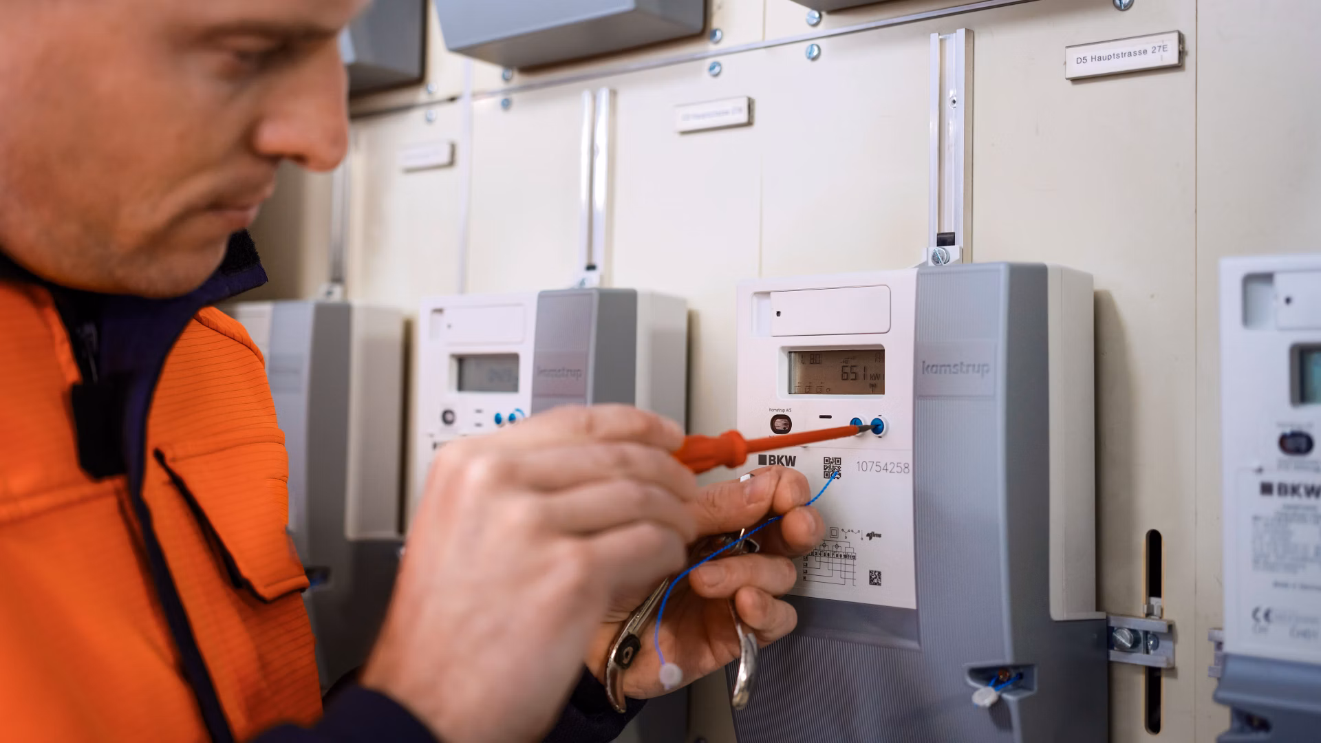 Technician in orange work clothing checks measuring devices in a control cabinet.