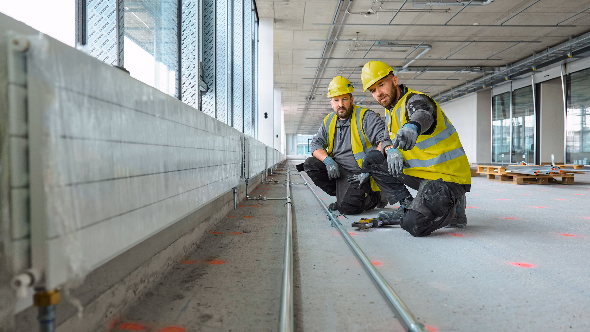 Zwei Bauarbeiter mit gelben Helmen und Warnwesten knien in einem Rohbau neben Leitungen am Boden.