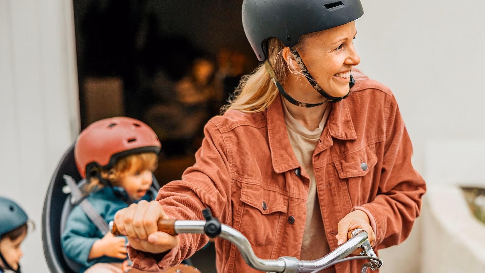 Frau mit Velohelm schiebt ein Fahrrad, während zwei Kinder mit Helmen daneben sitzen.